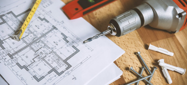 a drill, nails and a house plan on a wooden table to represent the first thing to do when you want to transform your basement into a livable space