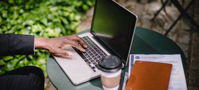 Mans hand typing on laptop keyboard, coffee and notebook next to laptop