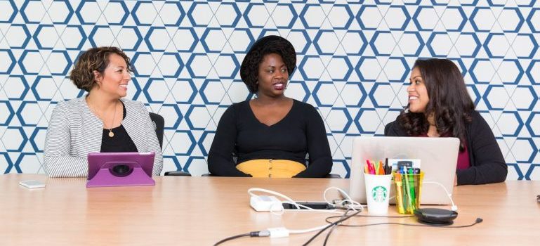 three women sitting in front of a wooden table and talking about how to help a friend move