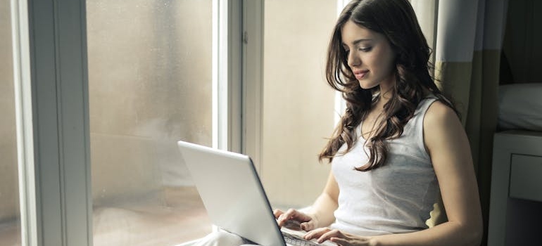 Woman sitting by the window and researching on her laptop.