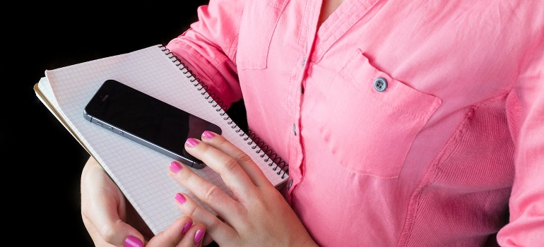 A woman in a pink shirt is holding a phone and a notebook.