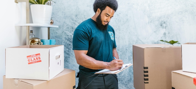 A Man in Blue Shirt Writing on a Clipboard Near Parcels